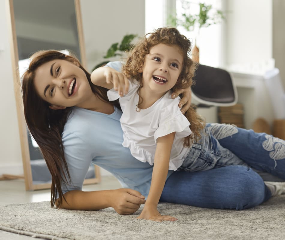 Mother and young daughter with curly hair laughing together on carpet in bright living room with wooden furniture and mirror.