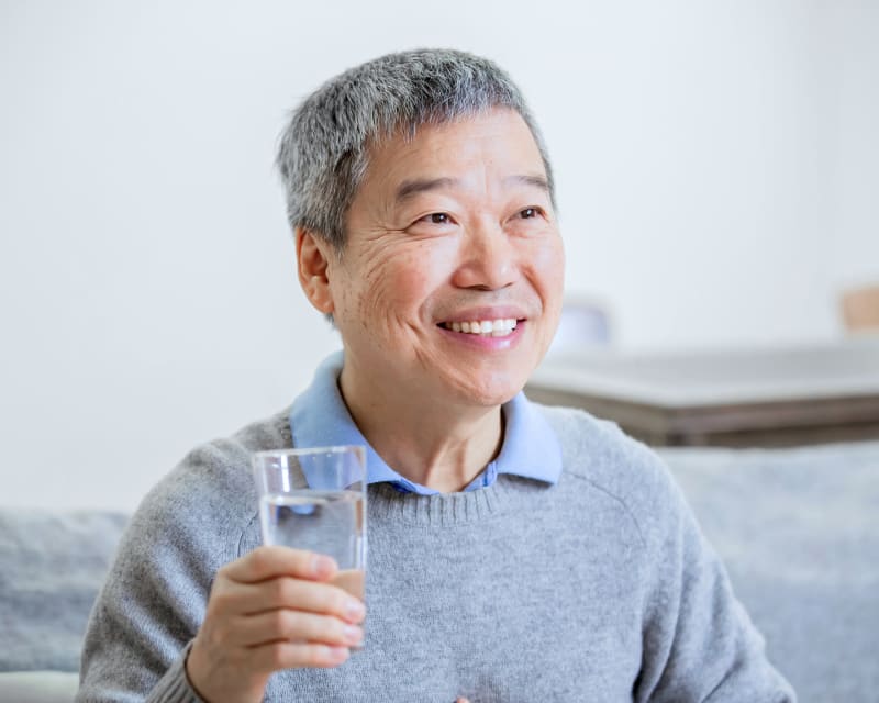 Older person with gray hair smiling while holding a glass of water, wearing a light blue shirt under a gray sweater.