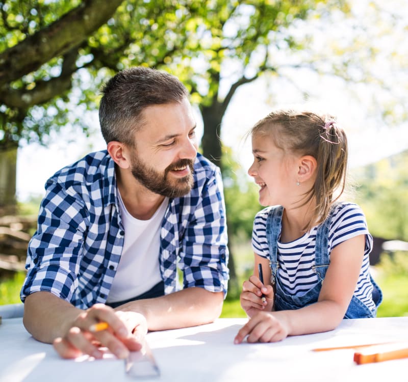 Bearded man in blue checkered shirt and young girl in striped shirt with denim overalls smiling at each other over white table with drawing supplies