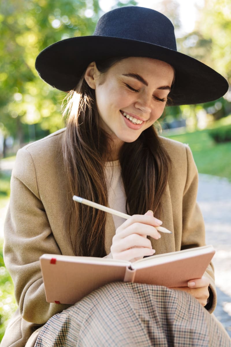 Young adult woman wearing a tan jacket and black hat is sitting in a park smiling while she writes in a journal.