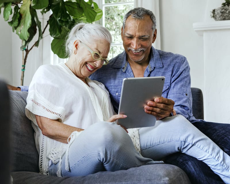 Older couple sitting on a soaf smiling and laughing while looking at a tablet with plants in the background.