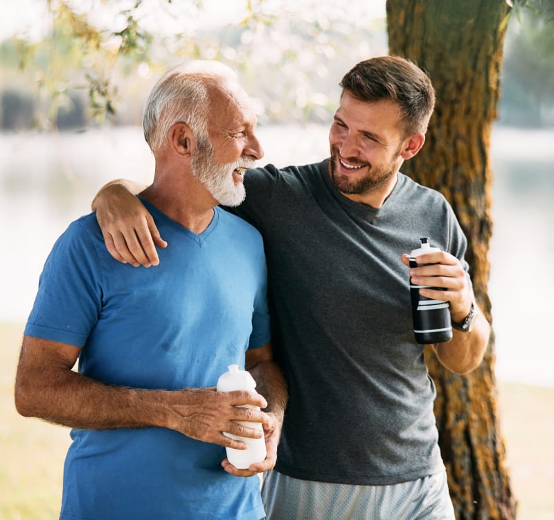 Older man in blue shirt and younger man in gray shirt sharing a laugh by a tree near a lake, both holding water bottles.
