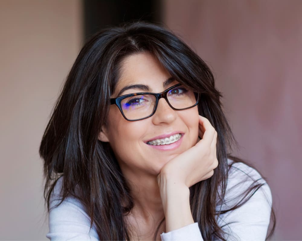 A woman with dark brown hair and glasses smiles at the camera, revealing a set of braces on her teeth.