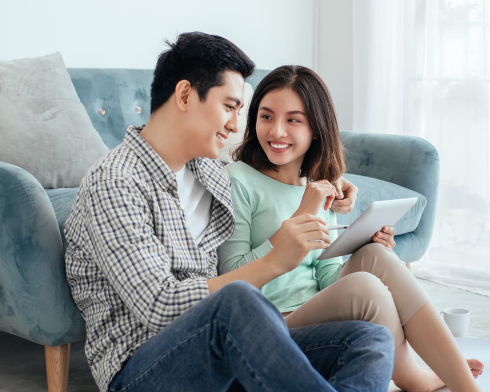 Couple sitting on floor in bright living room, smiling and looking at tablet with a blue sofa behind them.