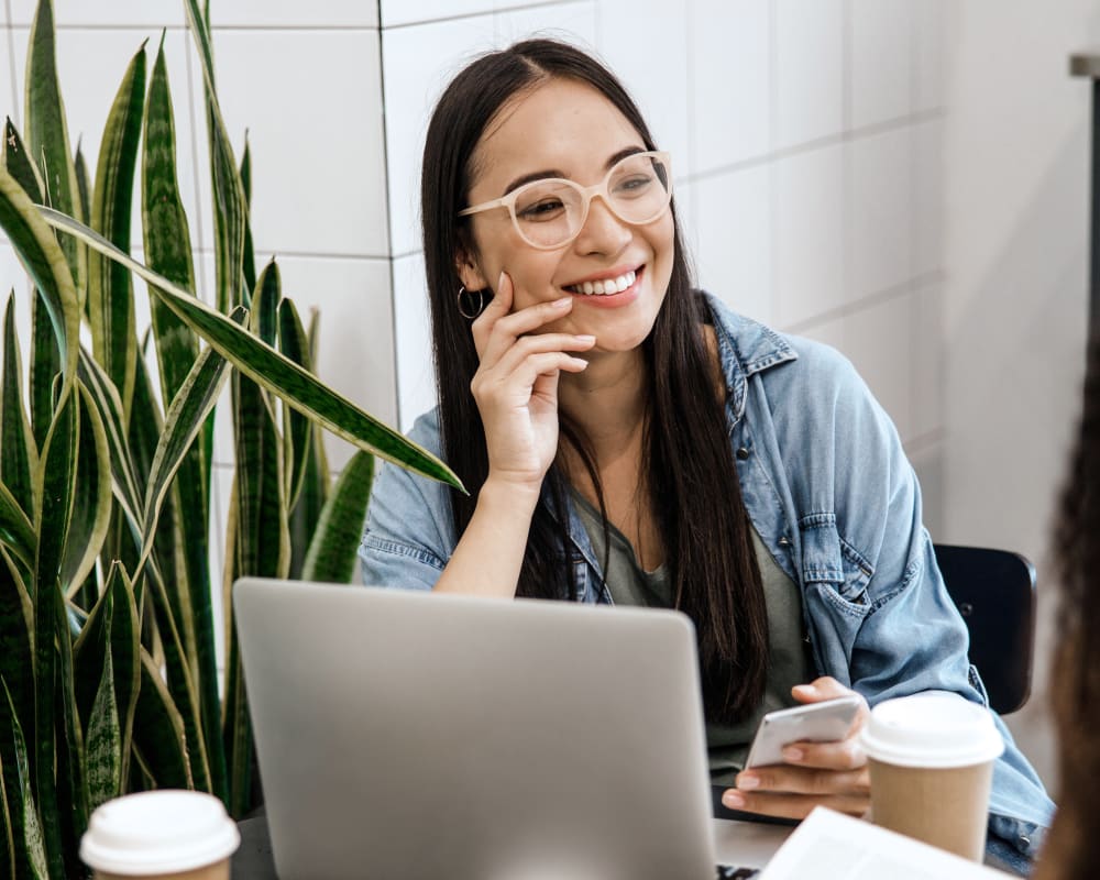 Young woman with long dark hair and white glasses smiling during a conversation at a cafe table with laptop, coffee cups, headphones and snake plants nearby.